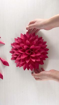 two hands reaching for a pink paper flower on a white surface with several petals scattered around it