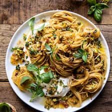 a white plate topped with pasta covered in sauce and herbs next to a glass of wine