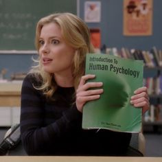 a woman sitting at a desk holding up a book in front of her face and looking off to the side