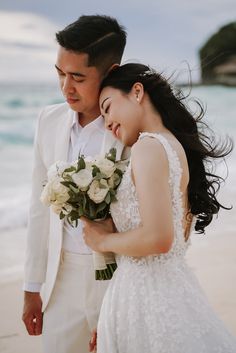 a bride and groom standing on the beach with their arms around each other holding flowers