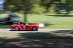 an old red sports car driving down the road in front of a truck and trees