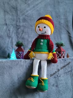 a crocheted snowman doll sitting on top of a gray blanket with green and yellow shoes