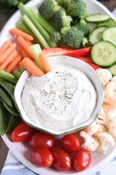 a white plate topped with veggies and dip surrounded by carrots, celery, cucumbers, tomatoes