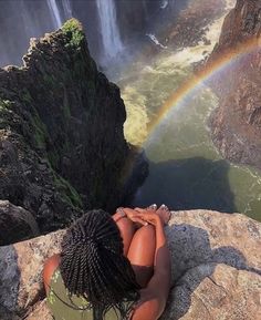 a woman sitting on top of a rock next to a waterfall with a rainbow in the background