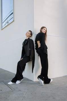 two women standing next to each other in front of a white wall and one is leaning against the wall