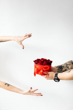 two hands reaching out towards each other holding a red box with roses in it and another person's arm behind them