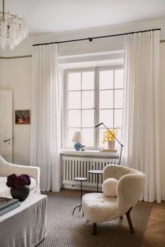 a living room with white curtains and a chair in front of a window that has a chandelier hanging from the ceiling