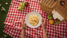 a person holding a plate with crackers and fruit on it next to a picnic blanket