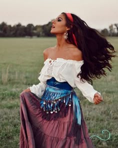 a woman with long hair wearing a skirt and white shirt standing in a grassy field