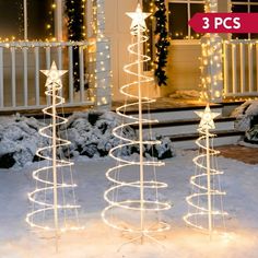 three lighted christmas trees in front of a house with snow on the ground and lights around them