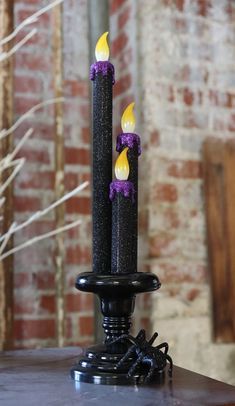 two black candles with yellow and purple lights on them sitting on a table in front of a brick wall