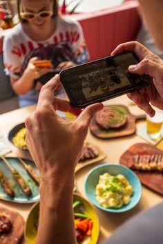 two people are sitting at a table with food and one person is holding up a cell phone