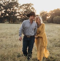 a man and woman are walking through the grass in an open field at sunset with trees in the background