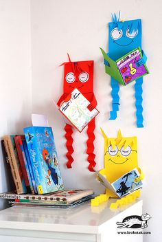 three paper monsters are hanging on the wall above a book shelf with books in front of them