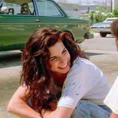 a man and woman sitting on the ground next to a green car with their arms around each other