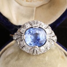 This exquisite ring was fashioned at the height of the Edwardian period. The masterfully crafted mounting is made in platinum and contains wonderfully rendered decoration: intricate engraving, filigree work, feminine ribbon motifs, a generous sprinkling of fine milgrain and 1.10ctw of old mine cut diamonds. A regal 4.25ct cushion cut periwinkle blue sapphire dazzles in the ring's central setting. This richly saturated, beautifully faceted blue stone hails from Sri Lanka (not so long ago known Edwardian Ring, Edwardian Jewelry, Traditional Diamond, Sapphire And Diamond Ring, Ceylon Sapphire, Antique Engagement, Antique Engagement Rings, White Jewelry, Periwinkle Blue