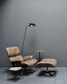 an eames chair and ottoman in front of a gray wall with a lamp on it