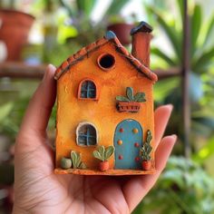 a hand holding a miniature house made out of clay with plants in pots behind it