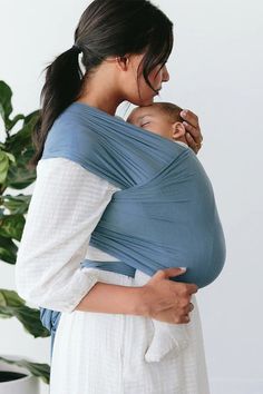 a woman holding a baby wrapped in a blue and white wrap on her back while standing next to a potted plant