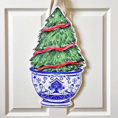 a christmas tree in a blue and white pot hanging from a door handle with ribbon on it