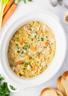 a white bowl filled with chicken noodle soup next to sliced carrots and bread