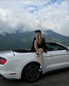 a woman sitting on the hood of a white convertible car with mountains in the background