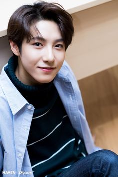 a young man sitting on the floor wearing a blue shirt and dark jeans, smiling at the camera