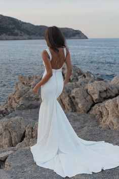 a woman in a white dress standing on rocks near the ocean with her back to the camera