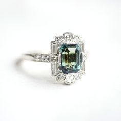 Item Details: 3.58 carat color change blue-green emerald cut sapphire Fourteen round brilliant cut diamonds 0.25 ct (G/VS) Two carre cut diamonds 0.14 ct (G/VS) Six baguette cut diamonds 0.36 ct (G/VS) Crafted in Platinum This ring was custom created with a one-of-a-kind center stone. Email custom@berlingerrings.com to create a similar ring. Enquire about this ring Design a custom piece with us Formal Emerald-cut Sapphire Ring With Halo Design, Heirloom Emerald-cut Sapphire Ring, Art Deco Emerald-cut Sapphire Anniversary Ring, Art Deco Engagement Ring Emerald, Green Emerald-cut Art Deco Diamond Ring, Luxury Art Deco Emerald-cut Sapphire Ring, Art Deco Sapphire Engagement Ring, Emerald Cut Sapphire Ring, Sapphire Engagement Ring Halo