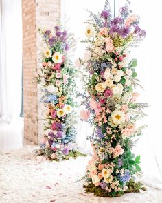 two tall floral arrangements on display in front of a brick wall with flowers all over them