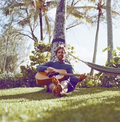 a man sitting on the grass with a guitar and hammock in front of him