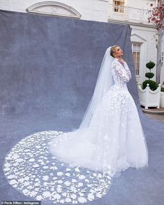 a woman standing in front of a backdrop wearing a white wedding dress with flowers on it