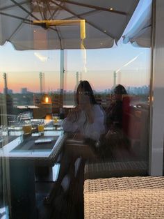 two women sitting at an outdoor table with umbrellas over their heads, looking out onto the city