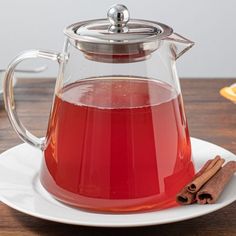 a glass tea pot filled with red liquid and cinnamon sticks on a white saucer