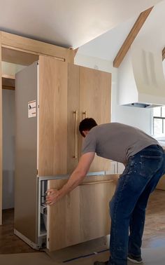 a man in grey shirt and blue jeans opening the door to a kitchen with wooden cabinets