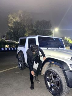 a man sitting on the tire of a white jeep parked in a parking lot at night