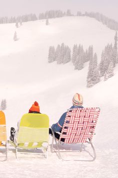 two people sitting in lawn chairs on top of a snow covered slope next to trees