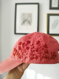 a person holding up a pink hat with red flowers on it in front of some framed pictures