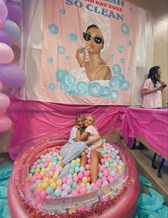 two people sitting in an inflatable pool with balloons and streamers on the floor