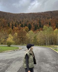 a woman standing in the middle of an empty road with trees on both sides and mountains in the background