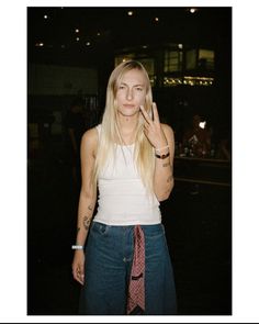 a woman with long blonde hair wearing a white tank top and blue jeans is holding her hand up to her face