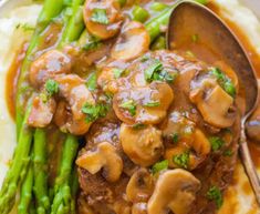 a white plate topped with mashed potatoes covered in mushrooms and asparagus next to a spoon