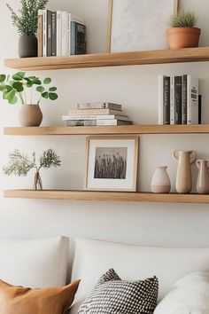 three wooden shelves above a couch with books and plants on top of them, in front of a white wall