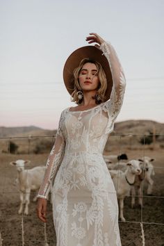 a woman in a white dress and brown hat standing next to a fence with cows