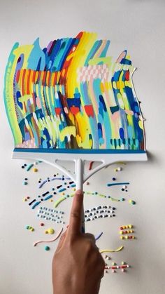 a hand pointing at an art piece on a white surface with multicolored paint sprinkles