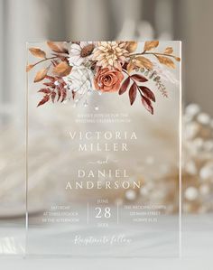 an acrylic wedding card with flowers and leaves is displayed on a white table