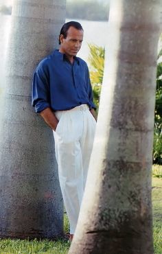 a man standing between two palm trees in front of some water and grass with his hands on his hips