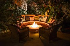 an outdoor fire pit surrounded by chairs and plants at night with lights shining on it