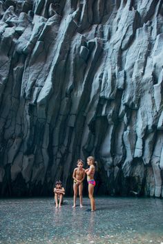 three people standing in the water next to a large rock formation