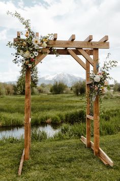 a wooden arch with flowers and greenery on the grass next to a pond in front of mountains